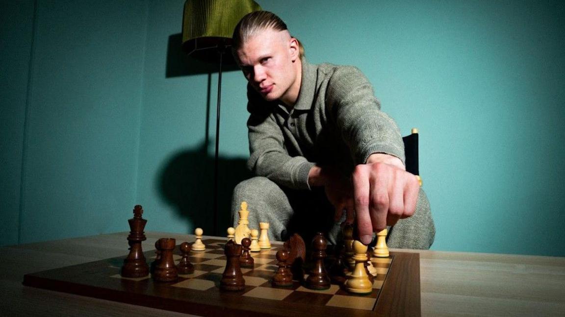 Erling Haaland looks into the camera as he moves a white bishop on a chess board.