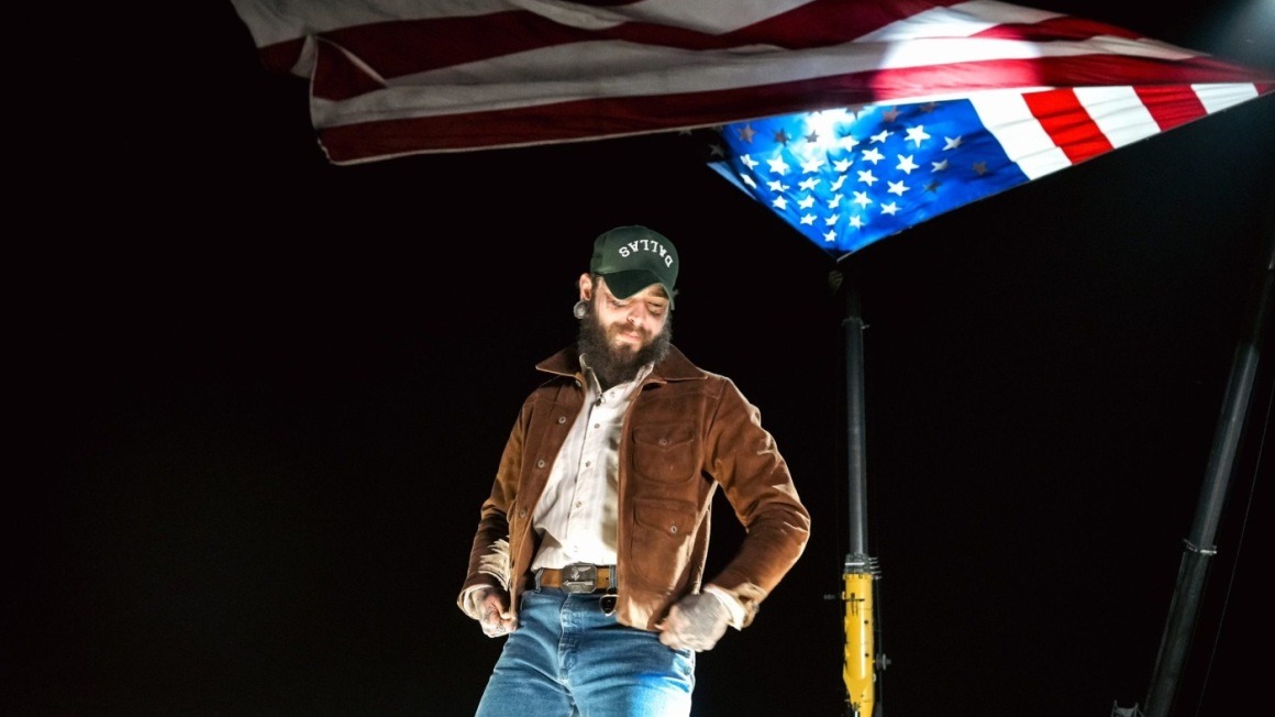 Post Malone wearing a cap and brown jacket standing in a pickup truck in a 2024 promotional photograph