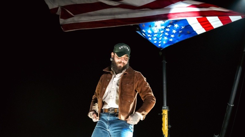 Post Malone wearing a cap and brown jacket standing in a pickup truck in a 2024 promotional photograph